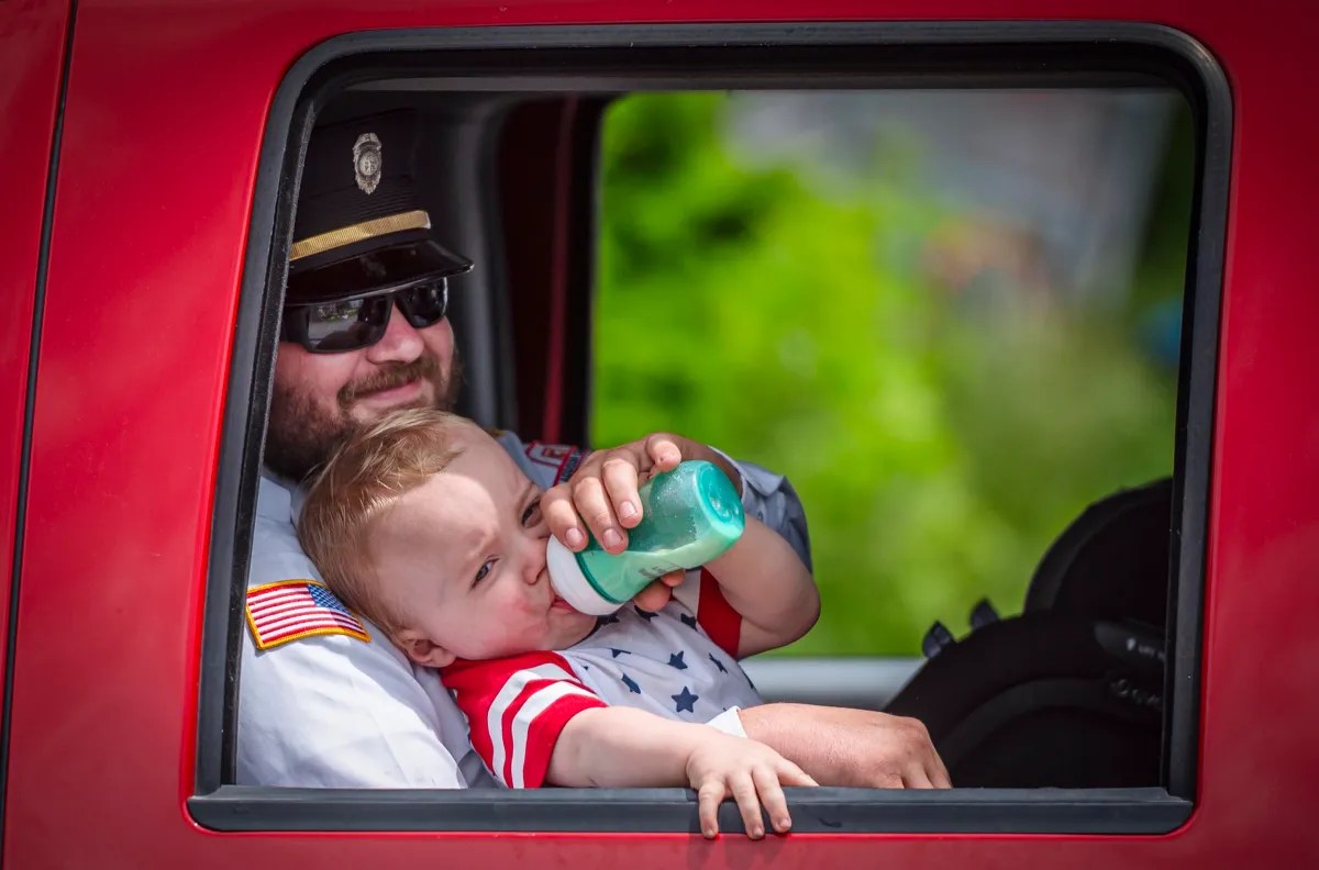 A man in uniform holds a baby drinking from a bottle inside a red vehicle.