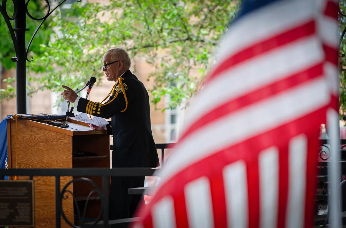 An individual in a formal uniform speaks at a podium outdoors, with an American flag in the foreground and green trees in the background.