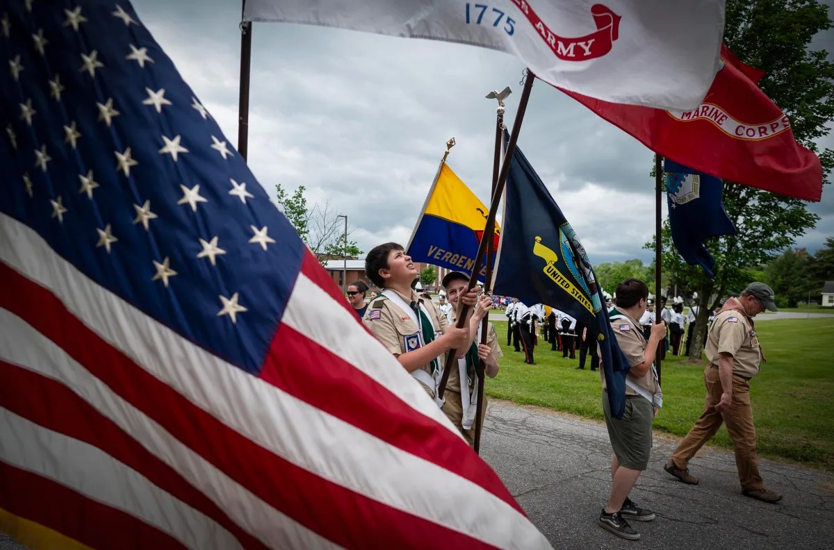 Boy Scouts holding various flags, including the U.S. flag, march in a parade. Other participants in uniform walk behind them on a cloudy day, with trees and grass in the background.