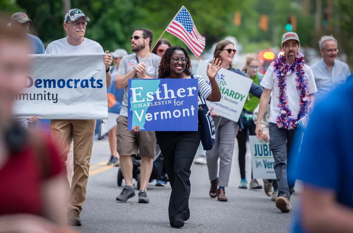 People participating in a political parade holding various campaign signs, including one for "Esther Charlestin for Vermont." An American flag is visible, and the participants seem engaged and cheerful.