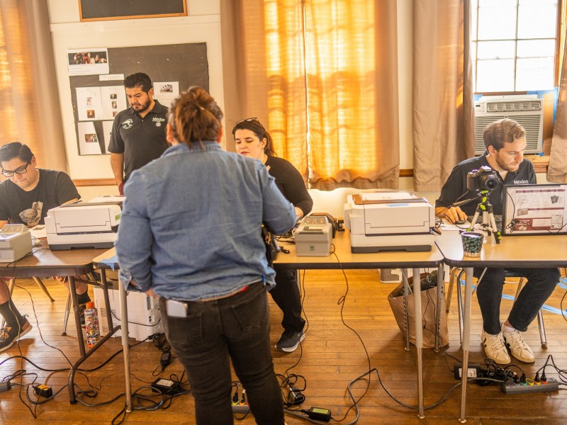Five people work at two tables with computers, printers, and various equipment in a room with wooden floors and large windows with tan curtains.