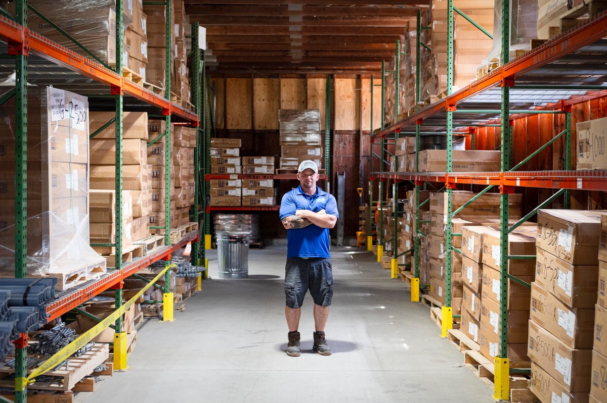 A person in a blue shirt and cap stands with arms crossed in the center aisle of a warehouse filled with stacked cardboard boxes and metal shelving.