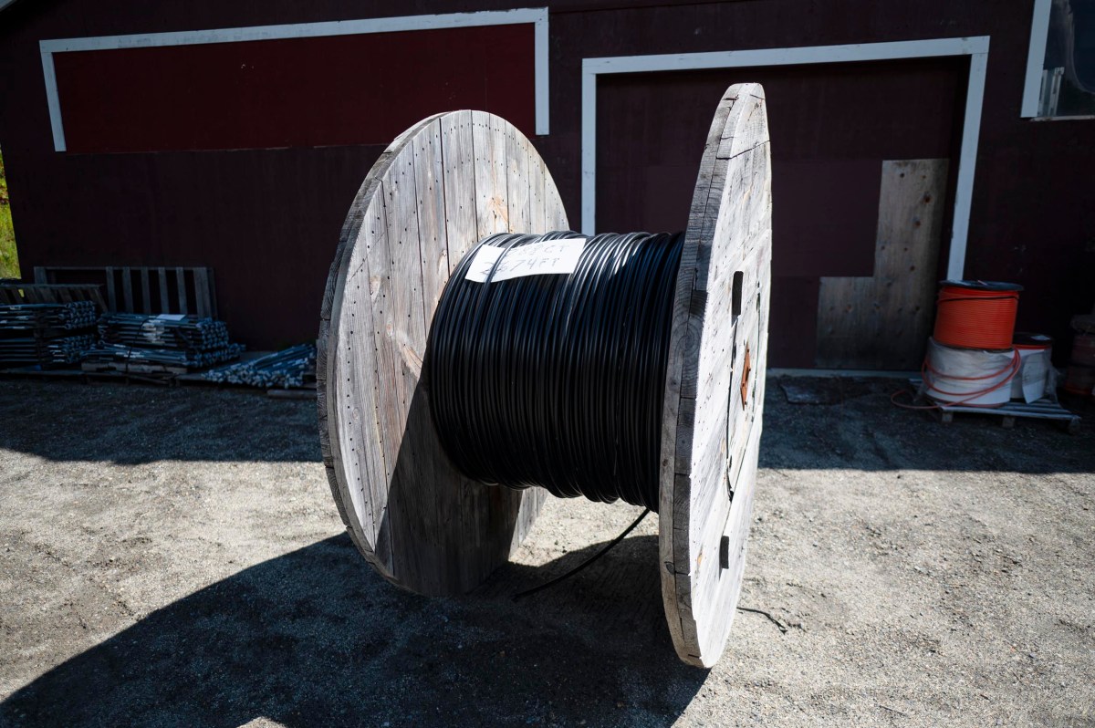 A large wooden spool with black cable is standing upright outdoors on gravel, in front of a dark red building.