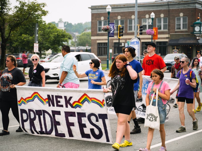 A group of people march in the street holding a banner that reads "NEK Pride Fest" with rainbow decorations during a parade.