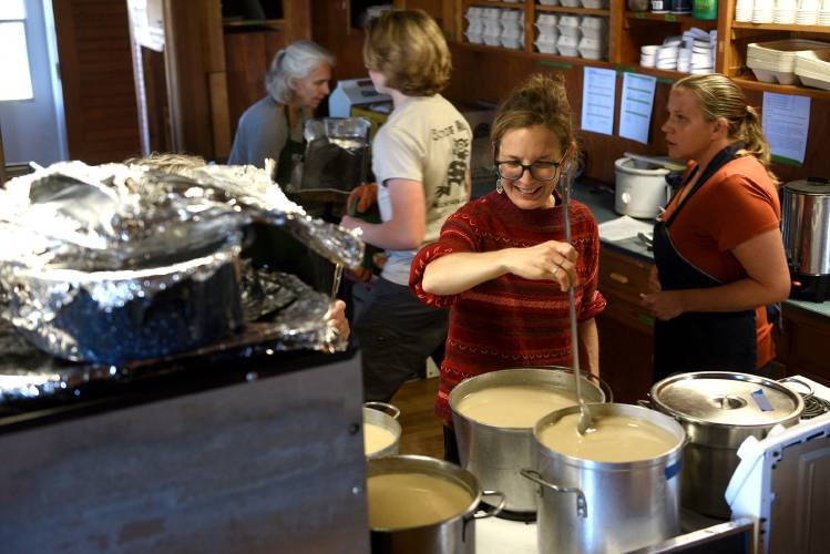 People in a kitchen ladling soup from large pots, with shelves of dishes in the background.