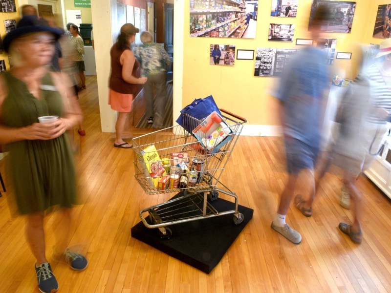 A shopping cart filled with groceries is displayed as an exhibit in an art gallery while people walk around it.