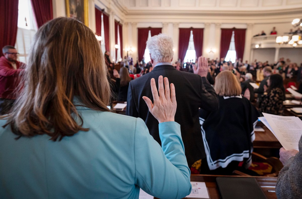 A group of people in formal attire stand with raised hands in a wood-paneled room, possibly taking an oath, facing a gathering with red curtains and chandeliers above.