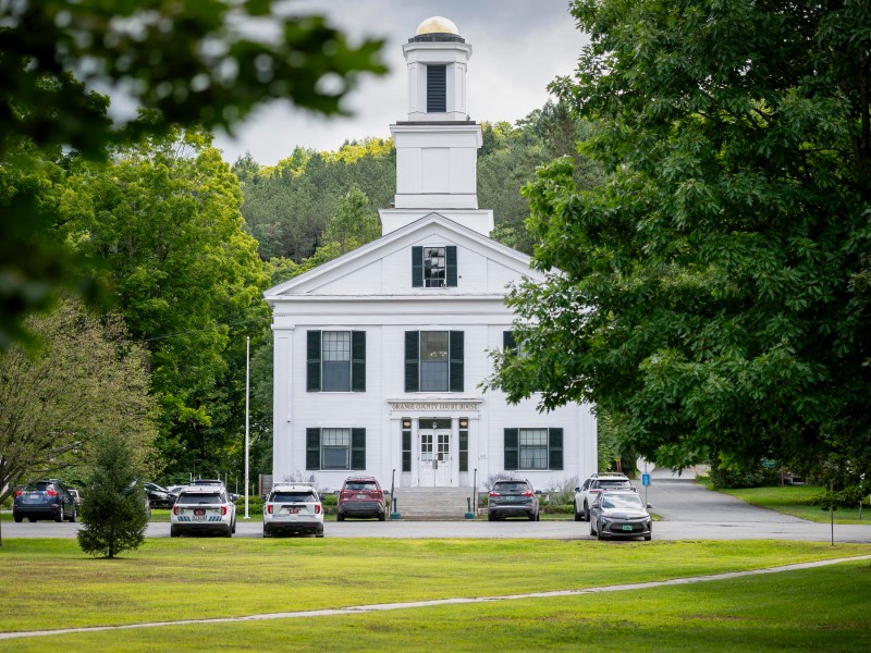 A historic white building with a tower, surrounded by trees and parked cars, stands on a green lawn under a cloudy sky.