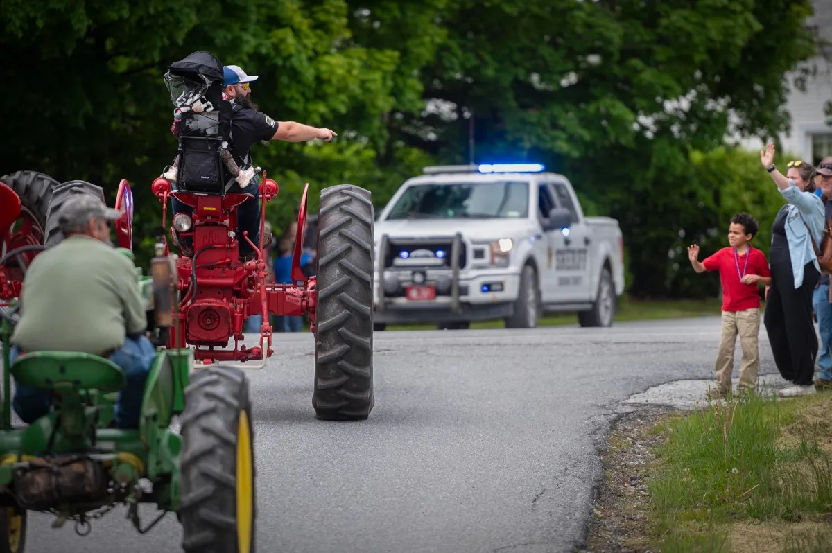 A person drives a red tractor past a police vehicle with flashing lights while people on the roadside wave and watch the parade.