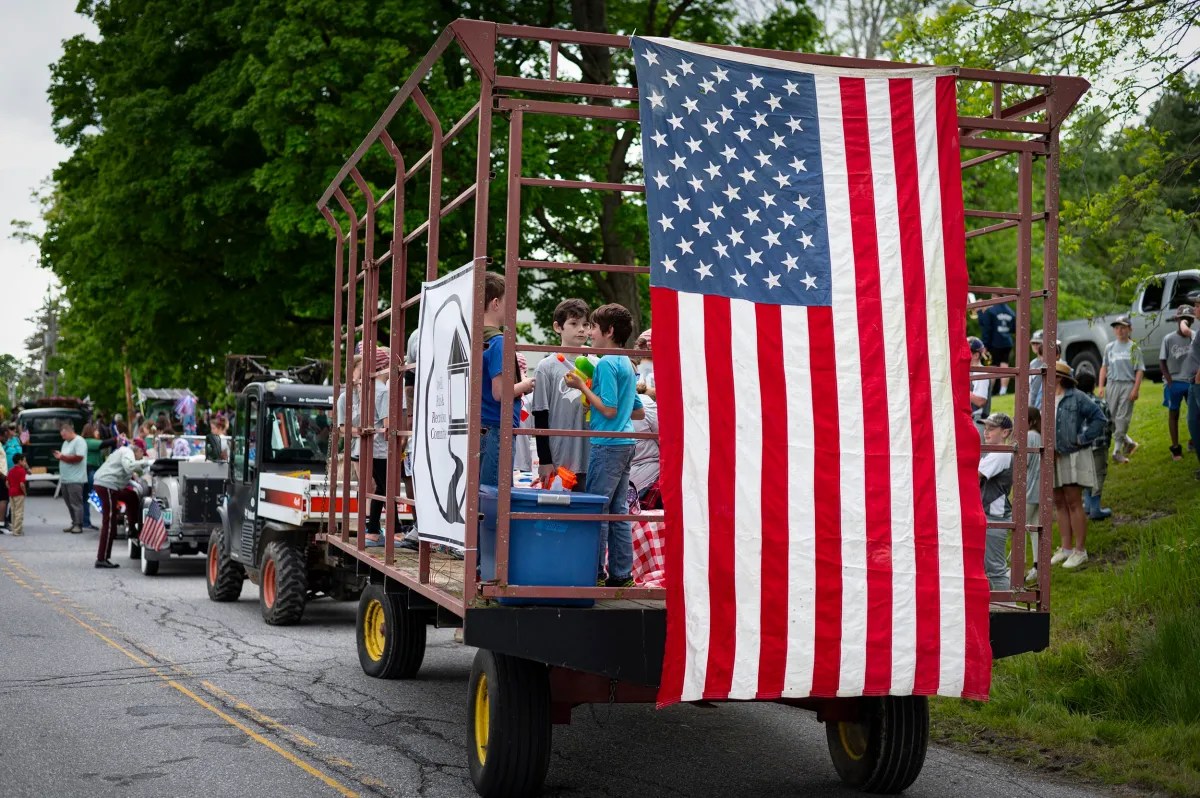 A group of children stand on a parade float decorated with a large American flag, moving along a street lined with bystanders and parked vehicles.