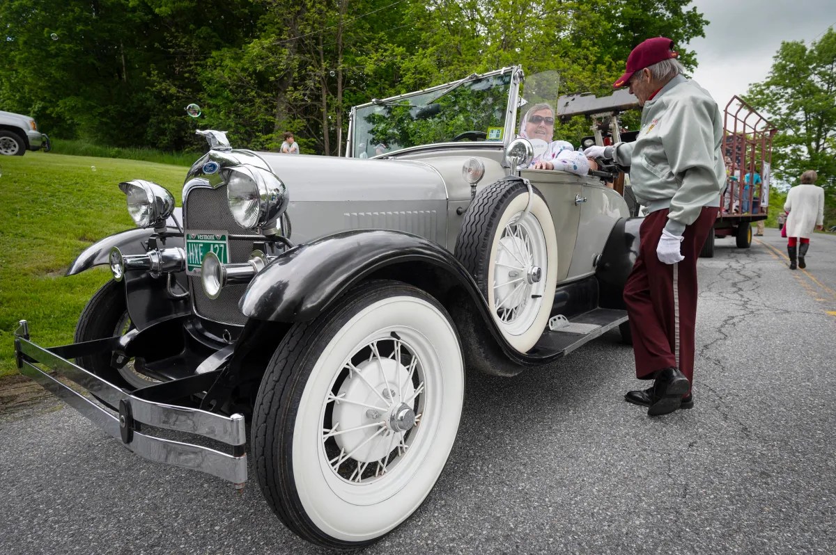 An elderly man in maroon pants stands beside a vintage convertible car parked on a road, with another person seated inside and trees in the background.