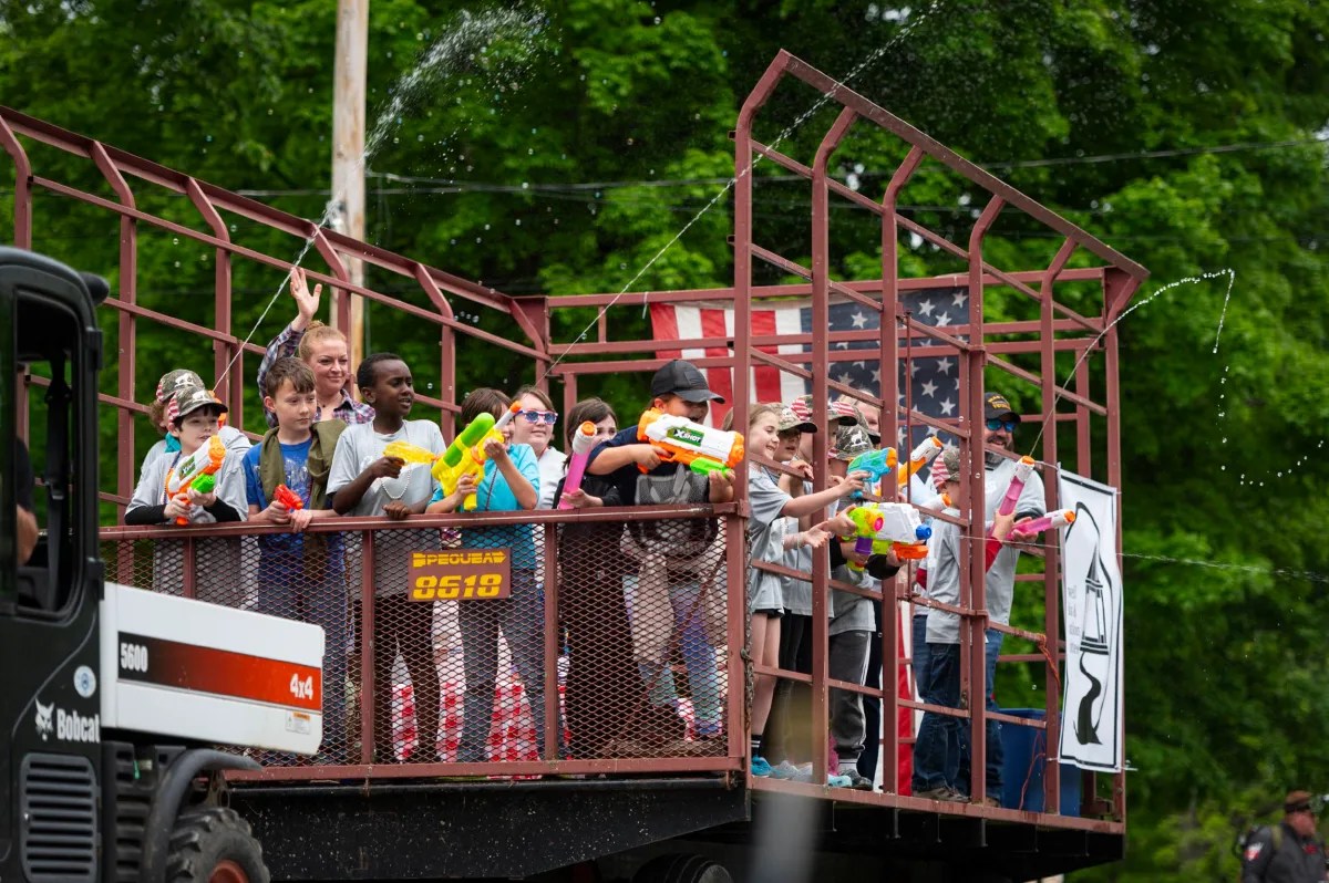 A group of children and adults stand on a fenced parade float, holding and spraying water guns, with trees and a flag in the background.