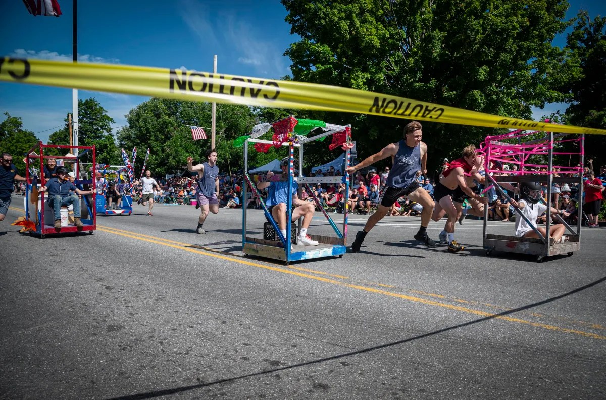 Participants push decorated carts in a road race during a community event, with a crowd of spectators in the background and a yellow caution tape in the foreground.