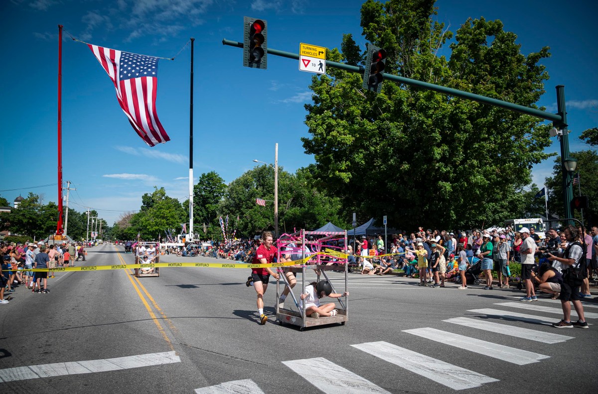 Participants pull a cart in a race on a street lined with spectators on both sides. An American flag is suspended above the road.