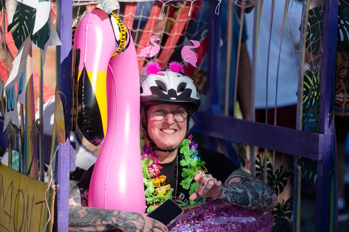 A person wearing a helmet decorated with fake flamingos smiles while holding a phone and standing next to a large inflatable flamingo. The scene is colorful with festive leis and banners.