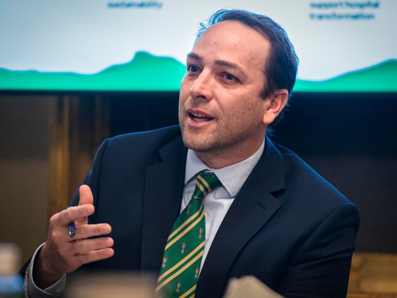 A man in a suit and tie is speaking in front of a screen.