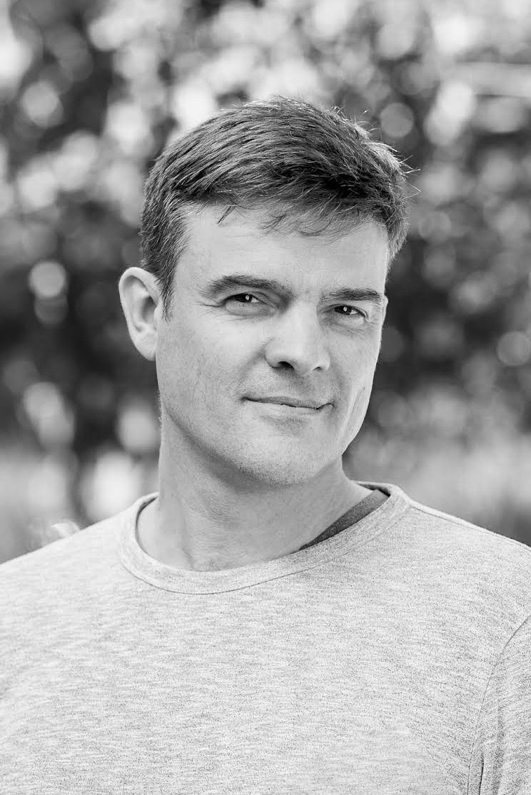 A man with short hair wearing a light-colored shirt is looking at the camera with a slight smile; background is blurred.