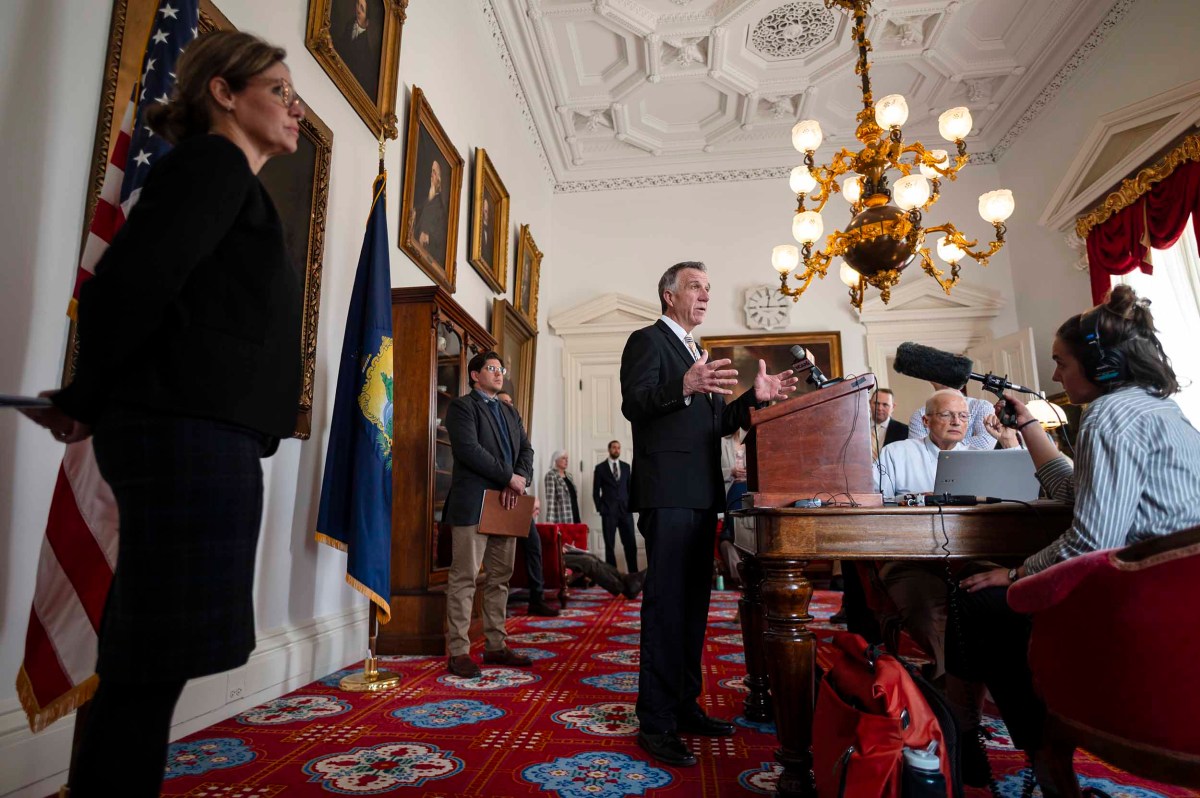 A man speaks at a podium in a white, ornate room with american flags, while people, including journalists with microphones, listen and observe.
