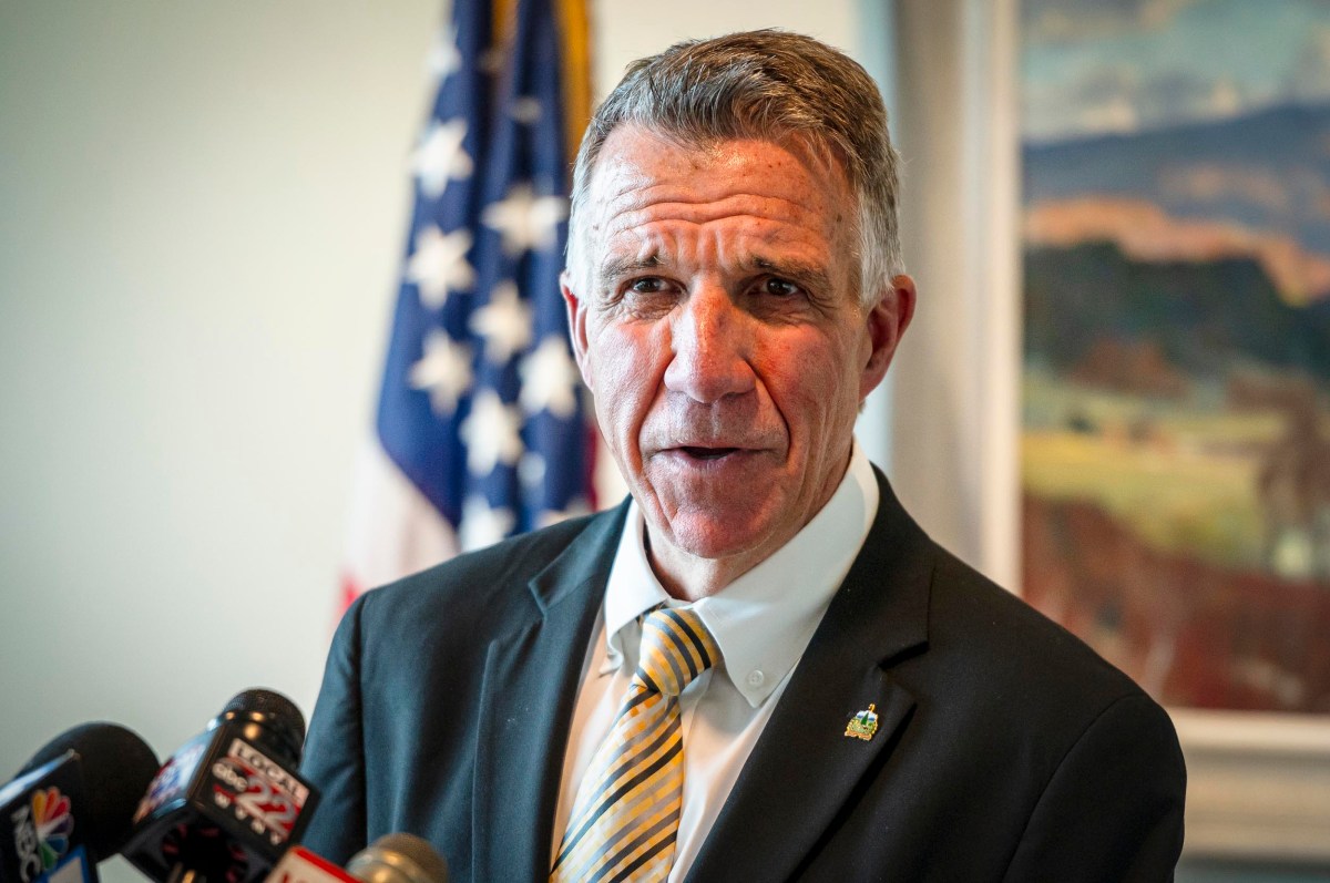 A man in a suit and striped tie speaks at a press conference in front of microphones. An American flag and scenic painting are in the background.