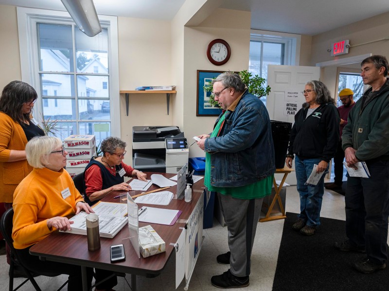 People are lined up at a table in a small room, one person is talking to a seated worker. There are posters on the walls and a clock showing 10:10.