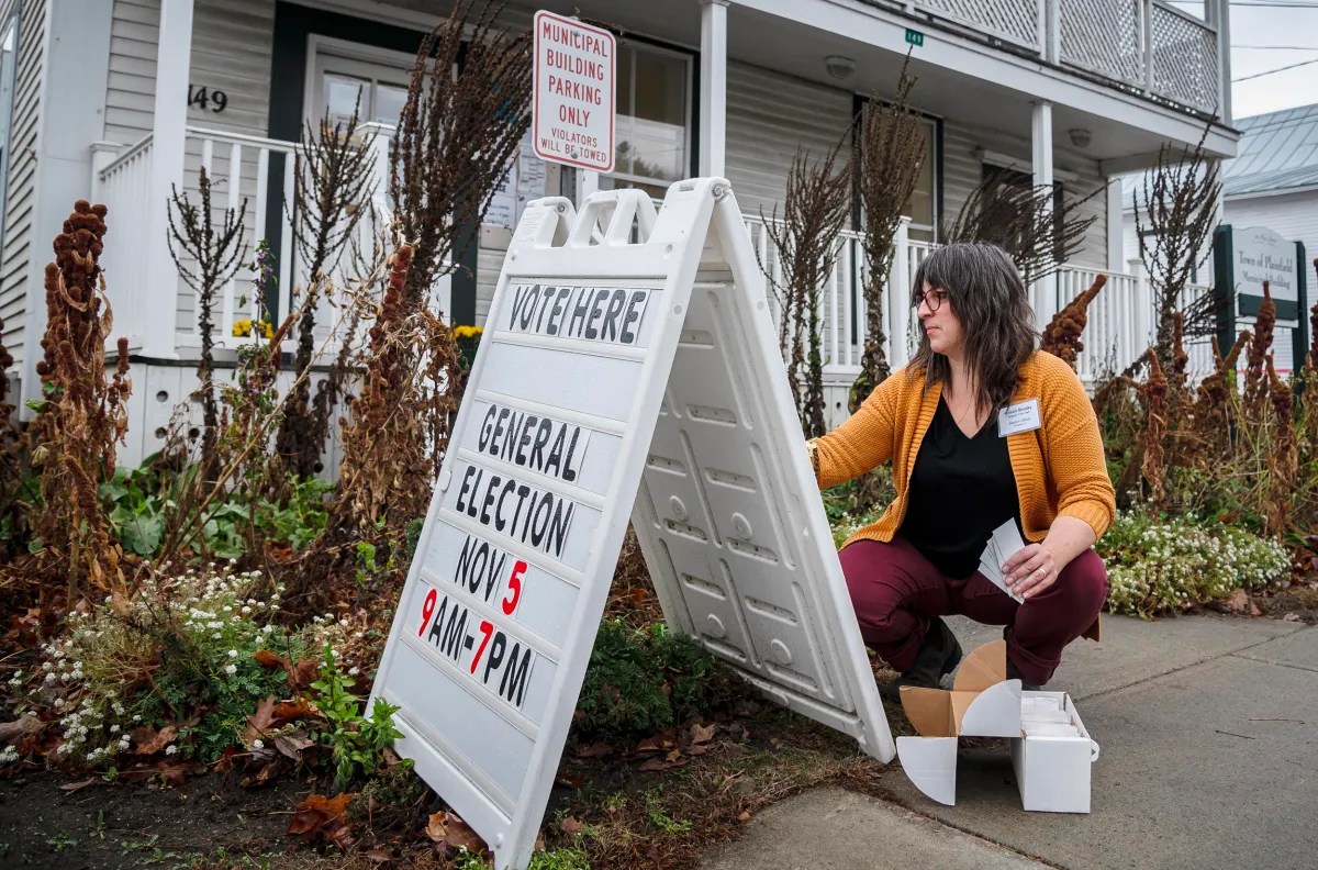 A woman sets up a "Vote Here" sign outside a building. The sign mentions a general election on November 5th, from 9 AM to 7 PM.