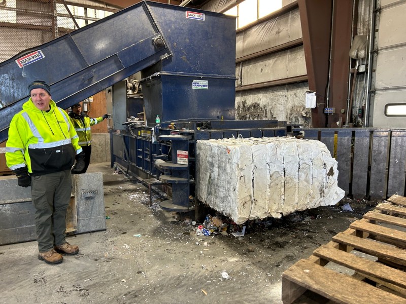 A worker stands next to a large recycling compactor machine compressing white paper material into a rectangular bale inside an industrial facility.