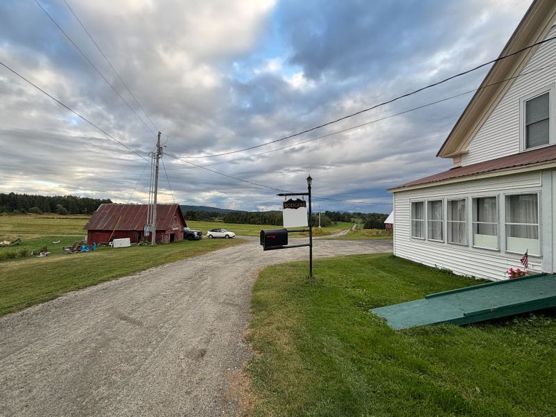 A gravel driveway leads past a white house and a red barn under a cloudy sky, with a truck parked nearby and fields in the background.