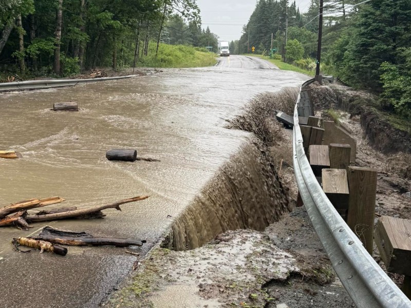 Floodwater flows over a damaged road, eroding the shoulder, with debris and logs scattered across the pavement, surrounded by trees.