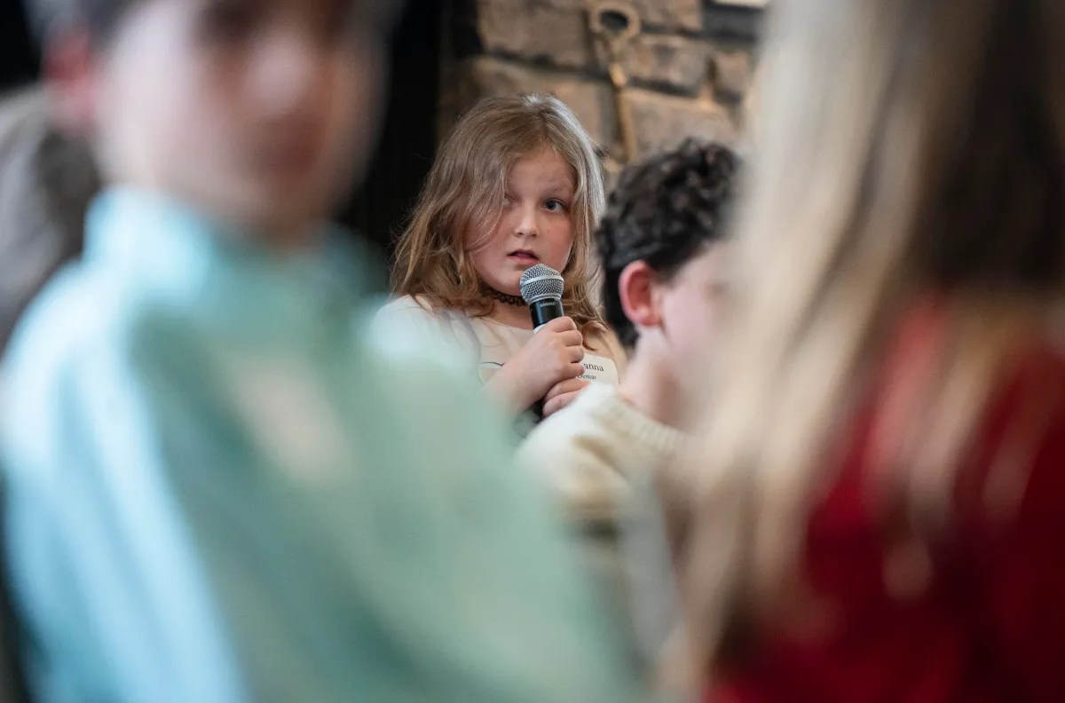 Young girl holding a microphone, looking pensive in a room with people.