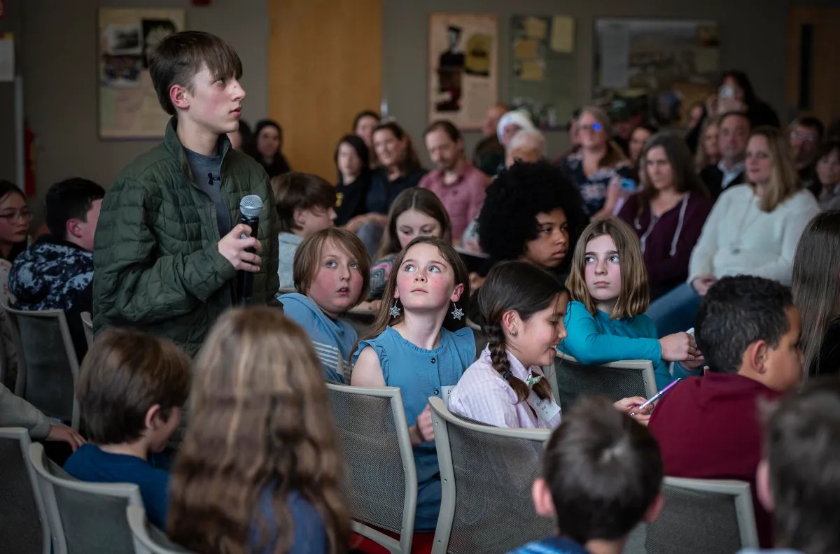 A young person holds a microphone and stands to address an audience, while children look on with attentive expressions.