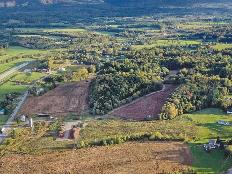Aerial view of a rural landscape with fields, roads, scattered houses, wooded areas, and distant mountains under a clear sky.