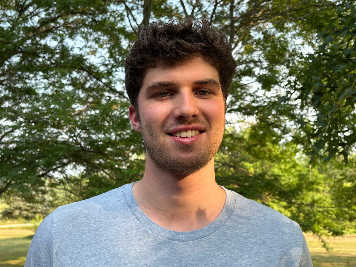 A young man with short, curly brown hair and a gray t-shirt smiles outdoors with trees and sunlight in the background.