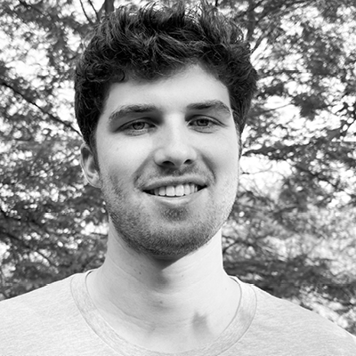 A young man with short curly hair and a light beard smiles outdoors, wearing a light-colored t-shirt with trees in the background.
