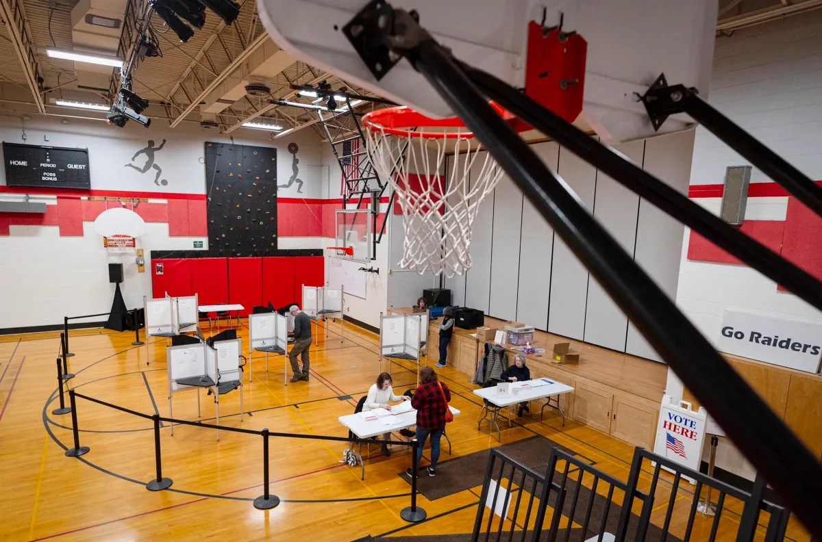 A gymnasium set up as a voting center with booths, tables, and signs. Voting equipment is spaced out, and a basketball hoop is visible in the foreground.