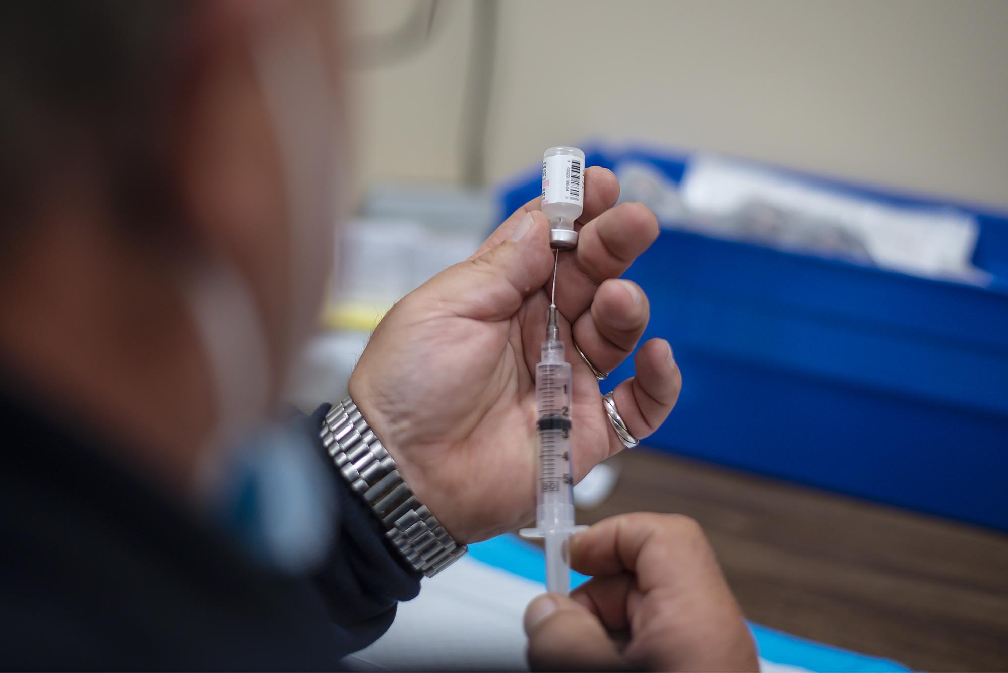 A person fills a syringe with liquid from a vial, preparing an injection. A blue tray and medical supplies are visible in the background.