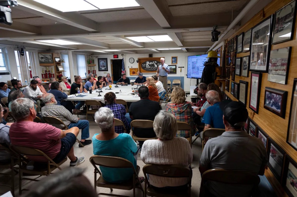 A group of people sit in a community meeting room, listening to a speaker presenting at the front near a screen displaying information.