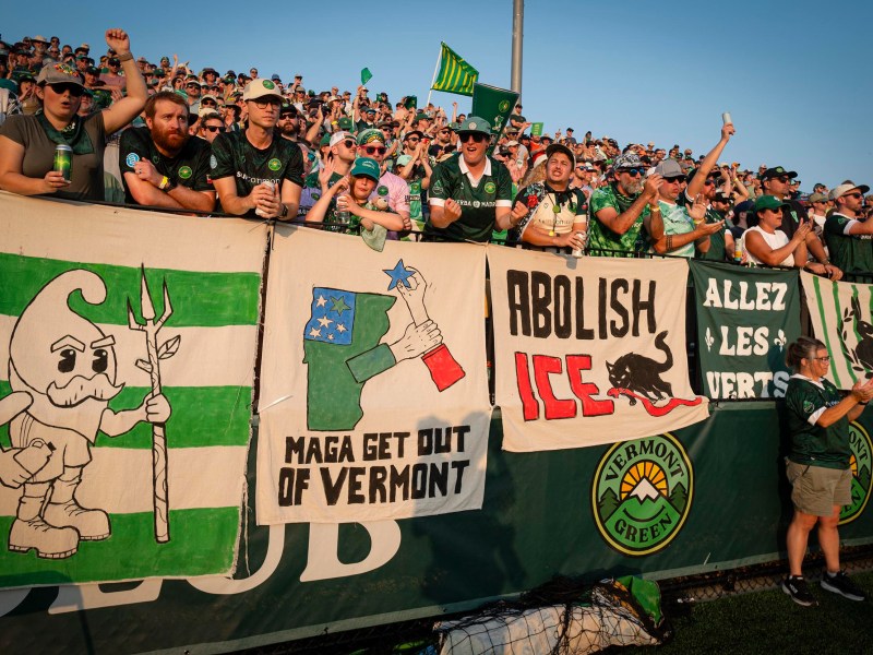 A crowd of soccer fans stands behind banners, including images and slogans such as “MAGA Get Out of Vermont,” “Abolish ICE,” and a Vermont Green FC logo.