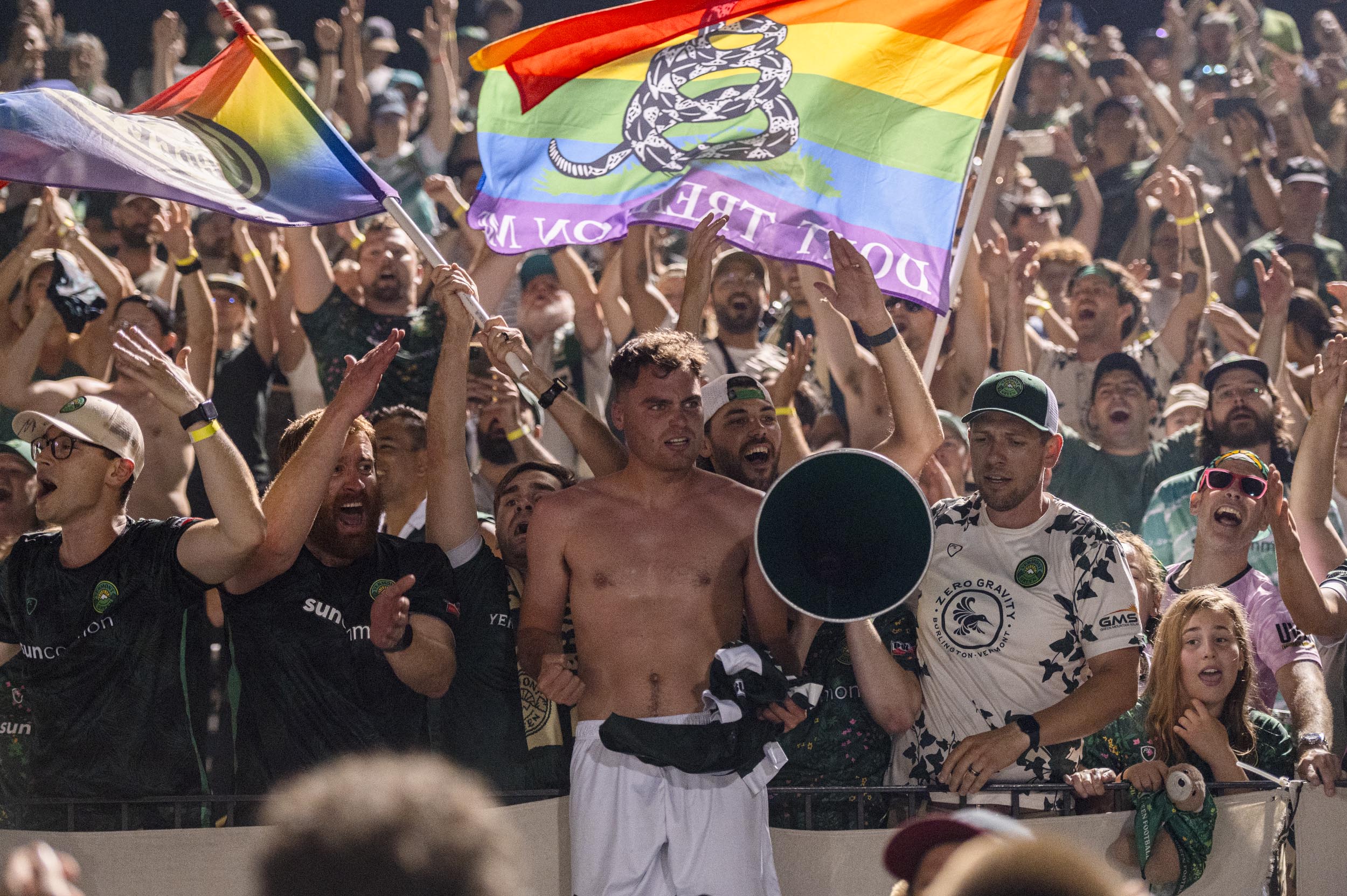 A group of cheering sports fans, some waving rainbow flags, surround a shirtless man holding a megaphone in a crowded stadium.