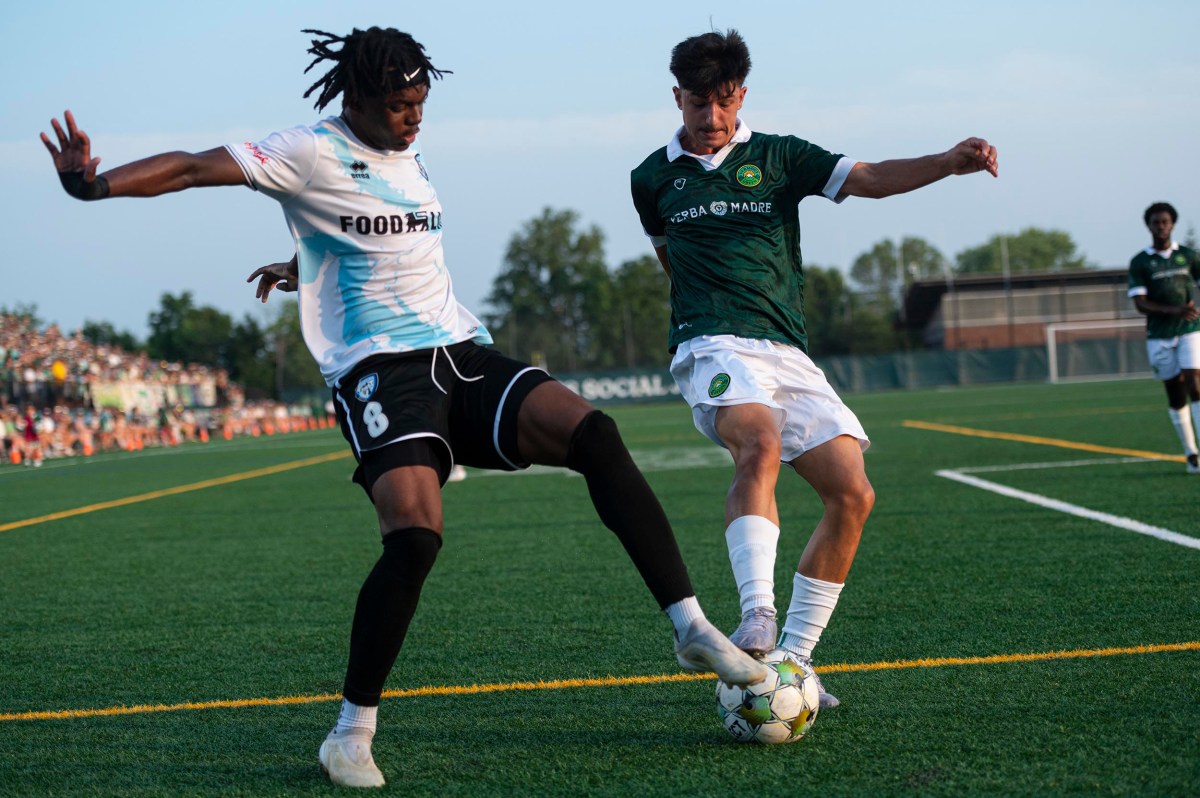 Two soccer players compete for the ball near the sideline during a match on a green field, with spectators visible in the background.