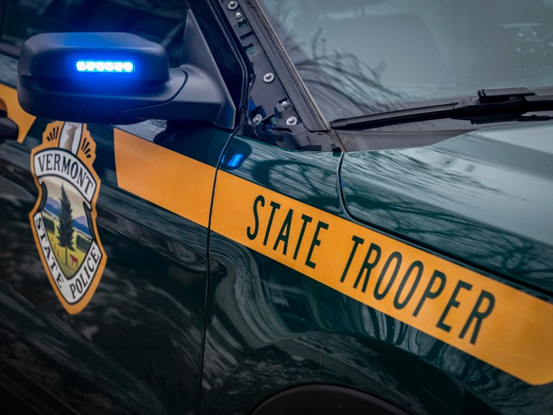 Vermont State Police vehicle with "State Trooper" text and emblem on the side, displaying a blue light on the mirror.
