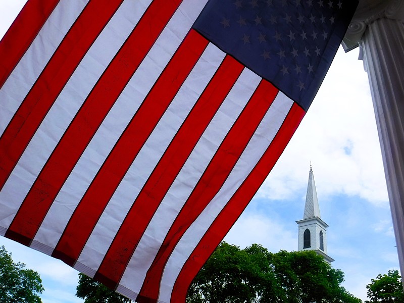 Large American flag in the foreground with a church steeple and trees in the background under a partly cloudy sky.
