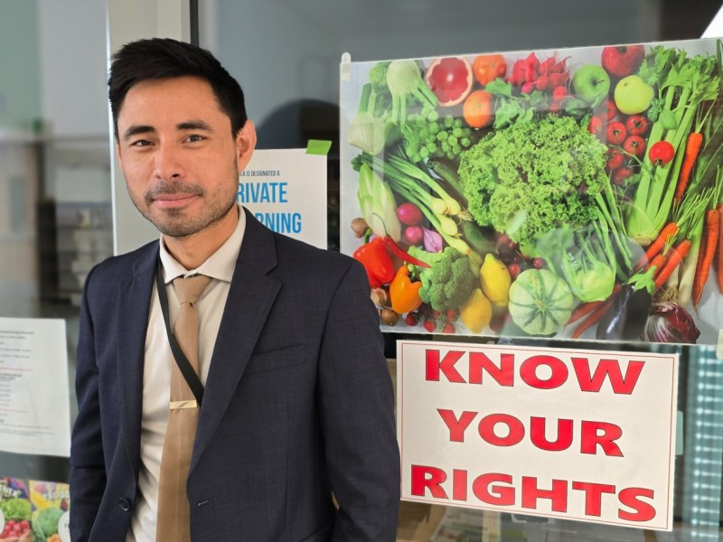 A man in a suit stands next to a door with a large poster of assorted vegetables and a sign that reads "KNOW YOUR RIGHTS.