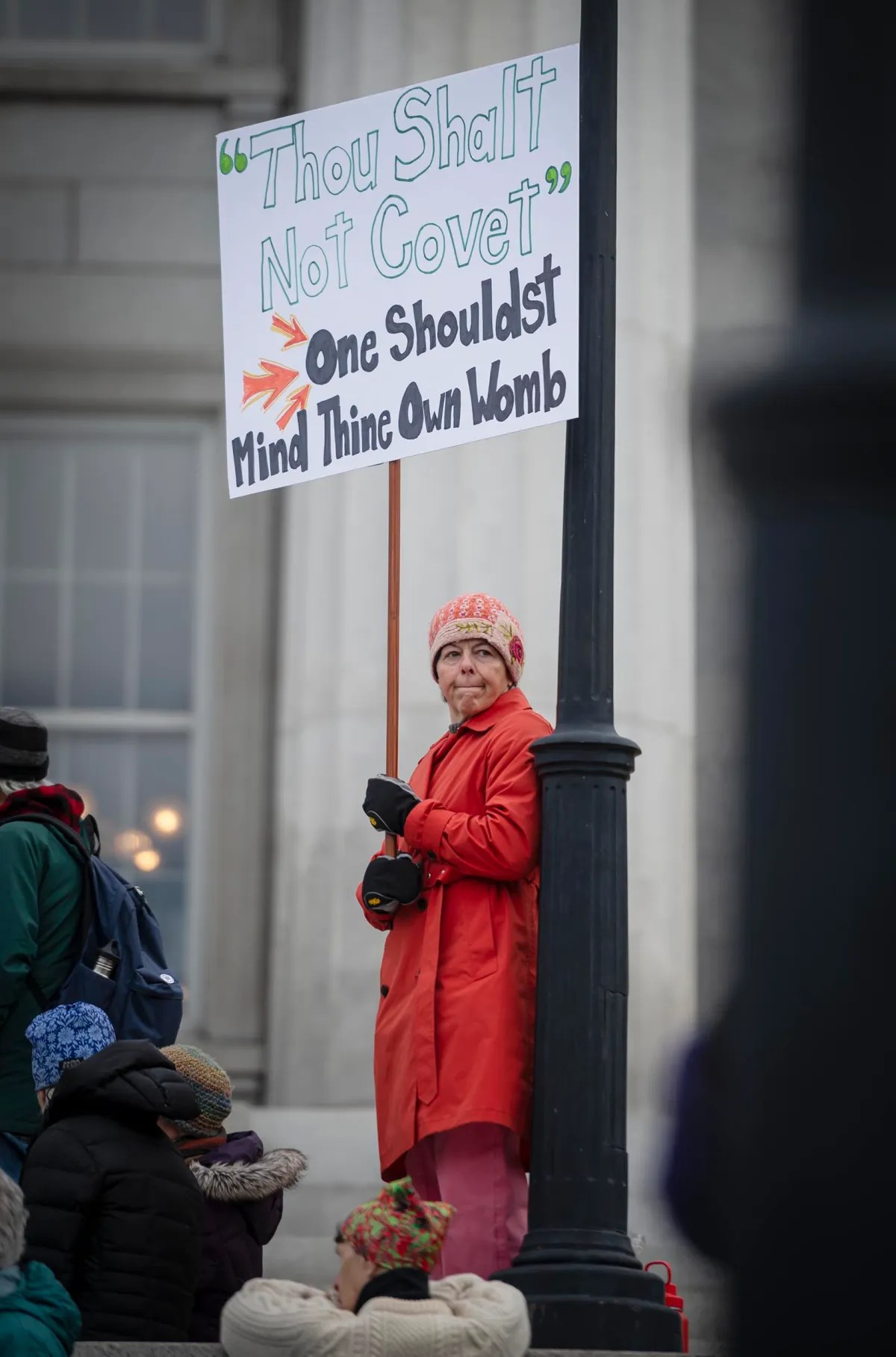 Person in a red coat holds a sign reading, "Thou Shalt Not Covet. One Shouldst Mind Thine Own Womb," while standing near a lamppost in a public area.