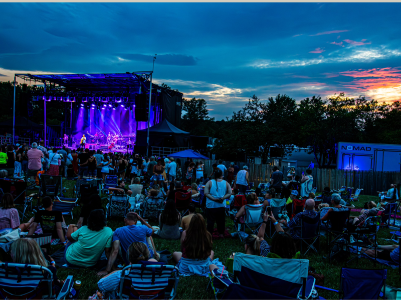 A crowd sits and stands on a grassy field watching a live outdoor concert on a stage at dusk, with colorful lights and a partly cloudy sky.